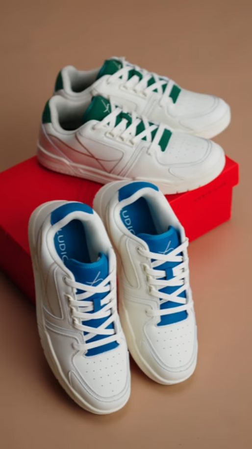 Two pairs of white sneakers with colored interiors and laces placed on and in front of a red shoebox on a beige surface.