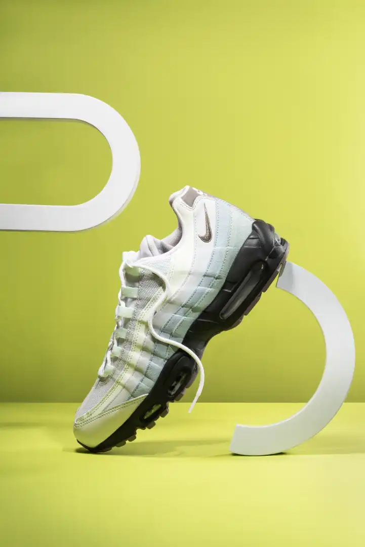 White and black Nike Air Max sneaker standing on a yellow surface with a light green background and white curved shapes.