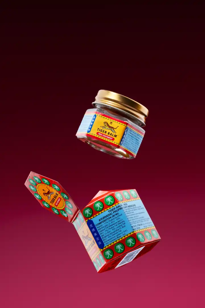 Open red and green box and small jar of Tiger Balm Red Ointment against a red gradient background.