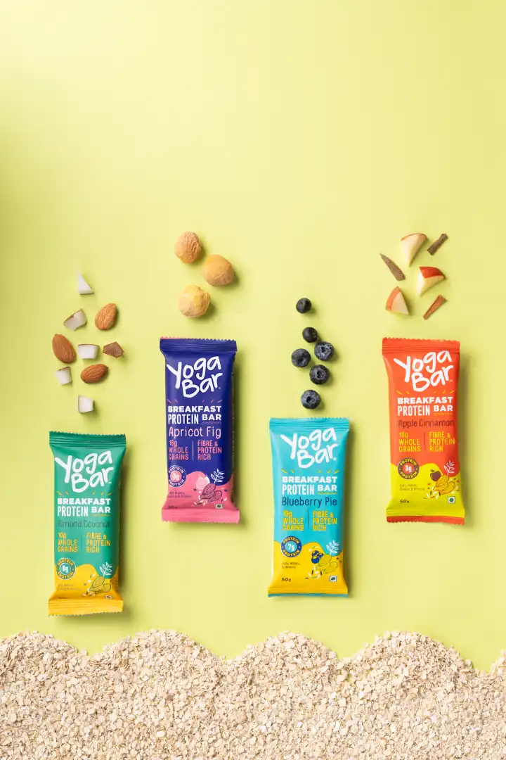 Four Yoga Bar breakfast protein bars in Almond Coconut, Apricot Fig, Blueberry Pie, and Apple Cinnamon flavors, each accompanied by their respective ingredients above them and oats at the bottom.