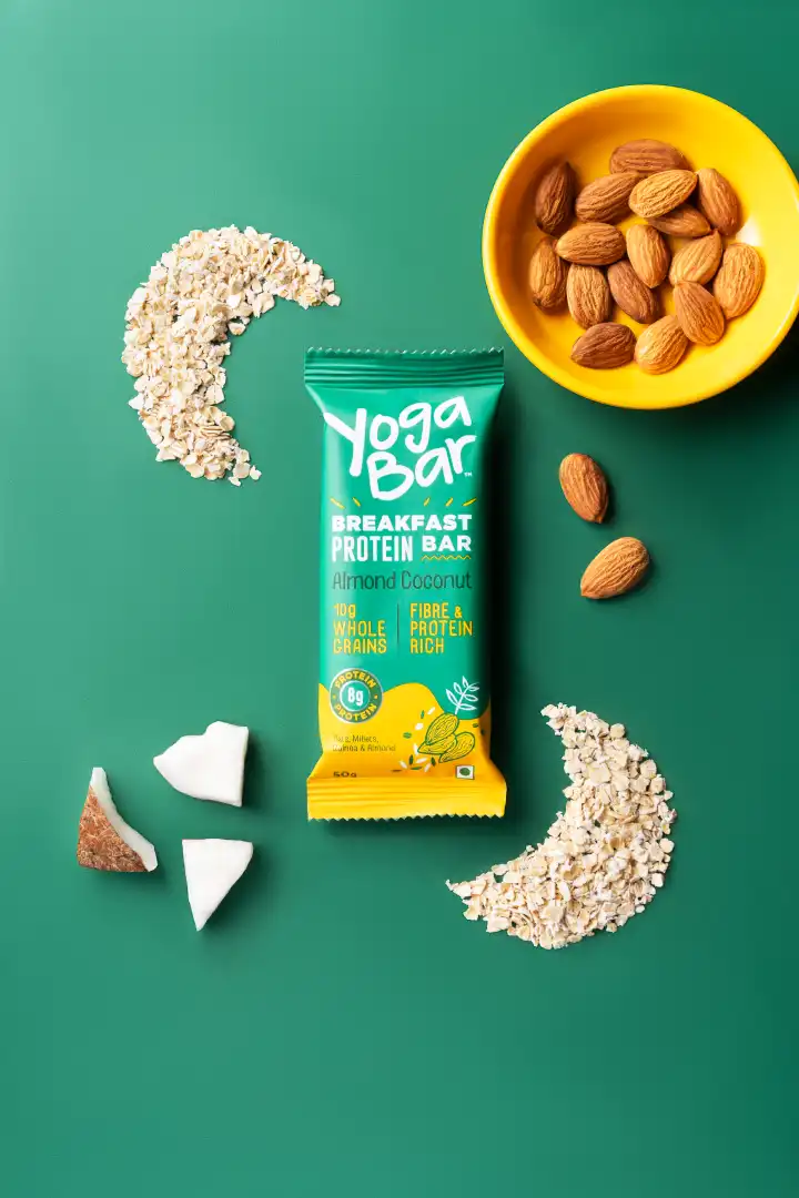 Yoga Bar Almond Coconut breakfast protein bar surrounded by oats, almonds in a yellow bowl, scattered almonds, and coconut pieces on a green background.