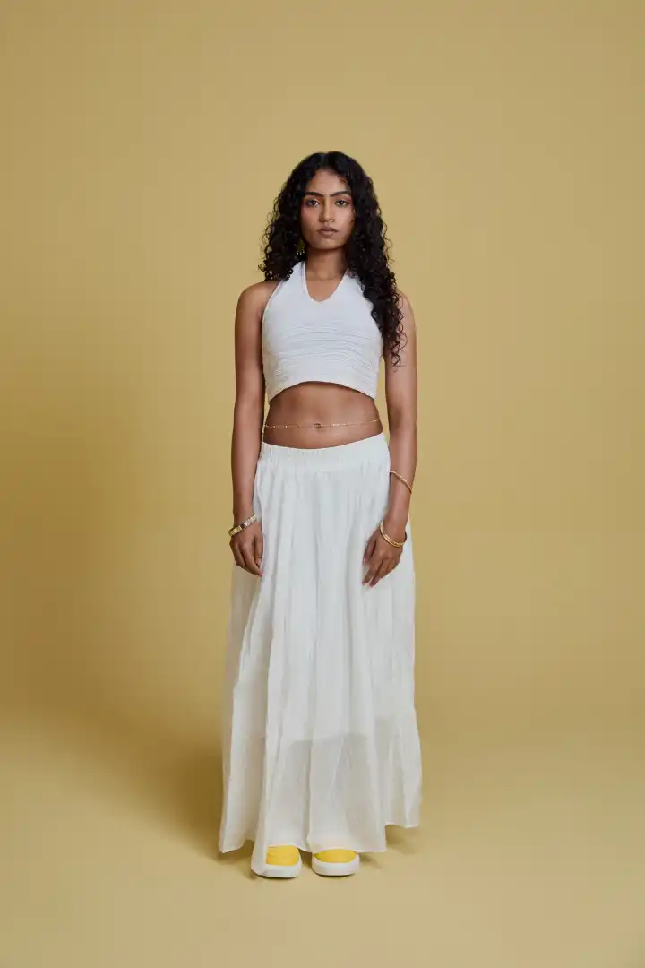 Woman with curly hair wearing a white sleeveless crop top, long white skirt, yellow sneakers, and gold jewelry, standing against a beige background.