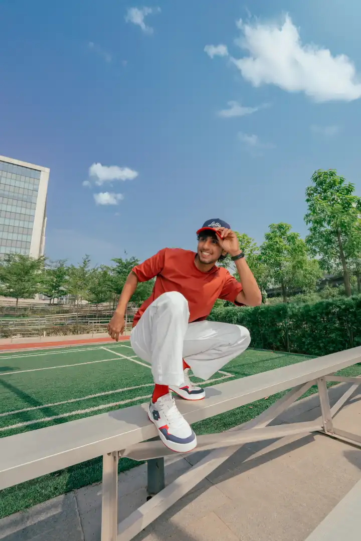 Smiling young man wearing a red shirt, white pants, and a cap crouching on an outdoor bench with a green field and trees in the background under a clear blue sky.