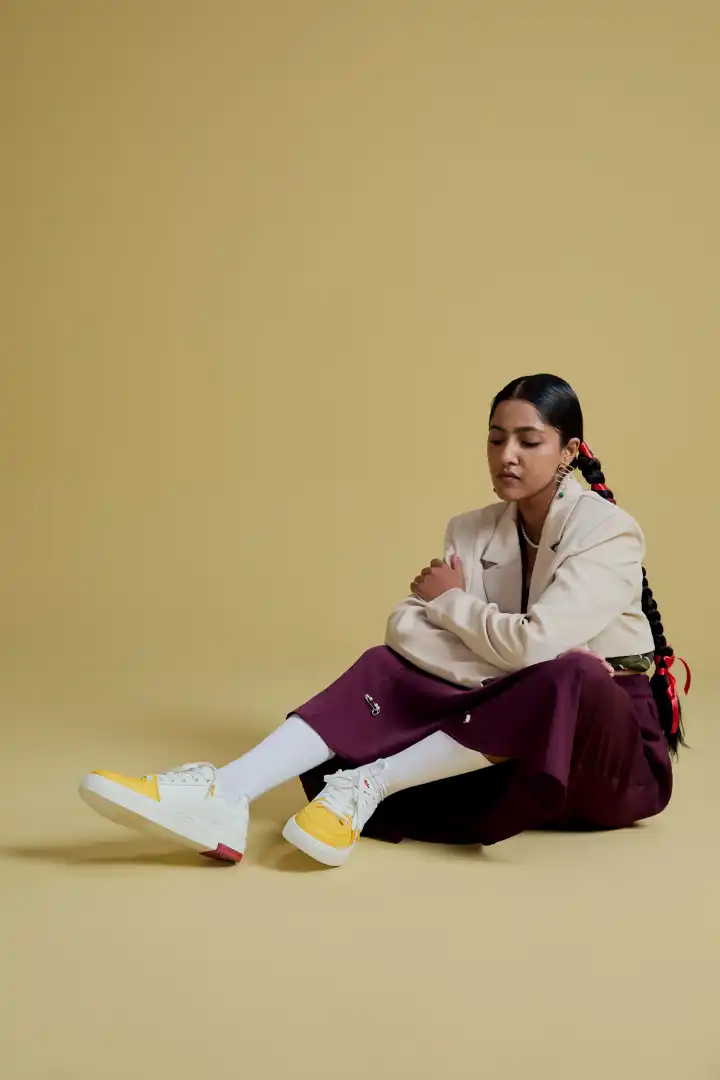 Young woman with long braided hair in a cream blazer, purple pants, white socks, and yellow-and-white sneakers sitting on a beige floor against a beige backdrop.