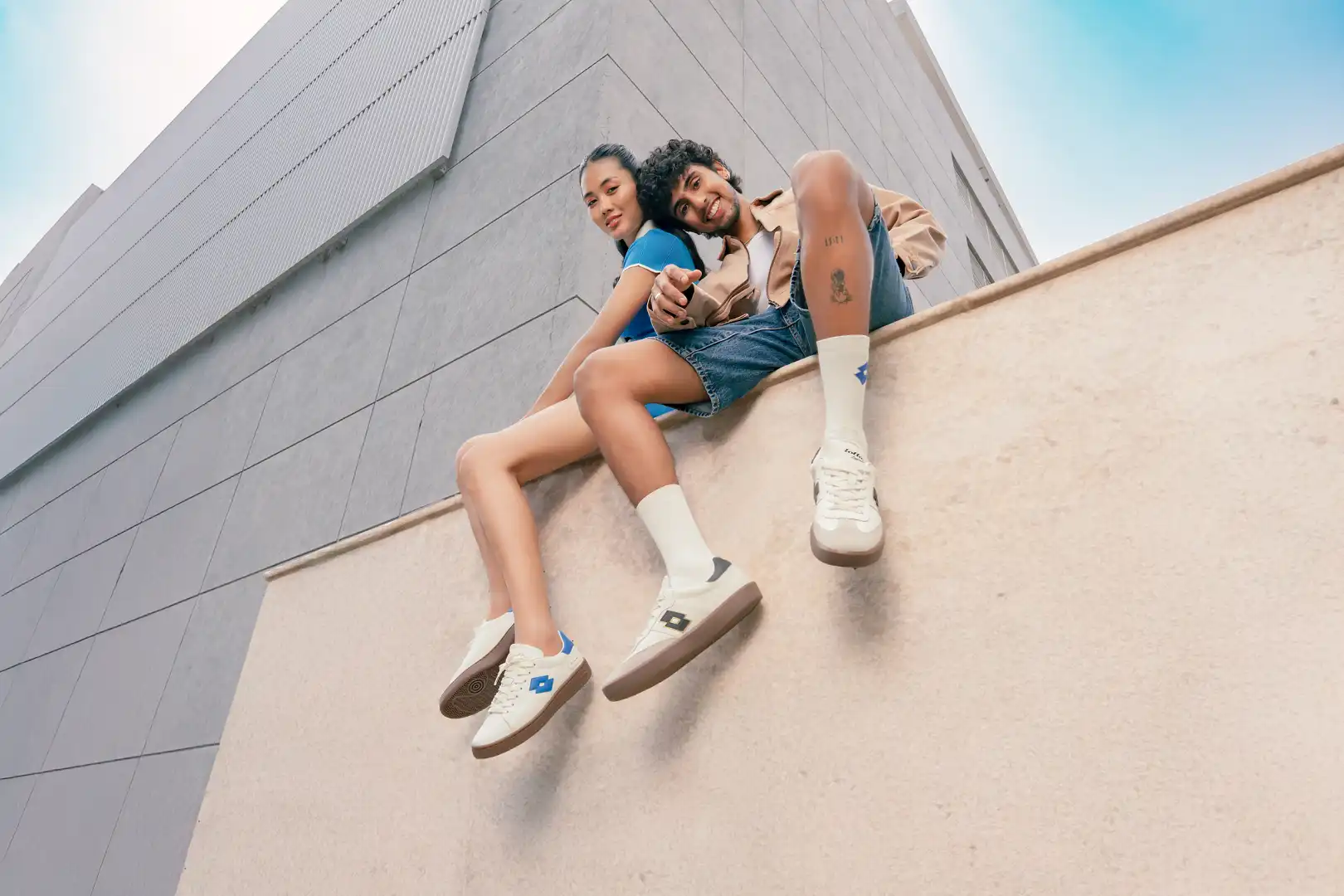 A young man and woman sitting on a ledge against a modern building, both wearing casual white sneakers with blue logos.