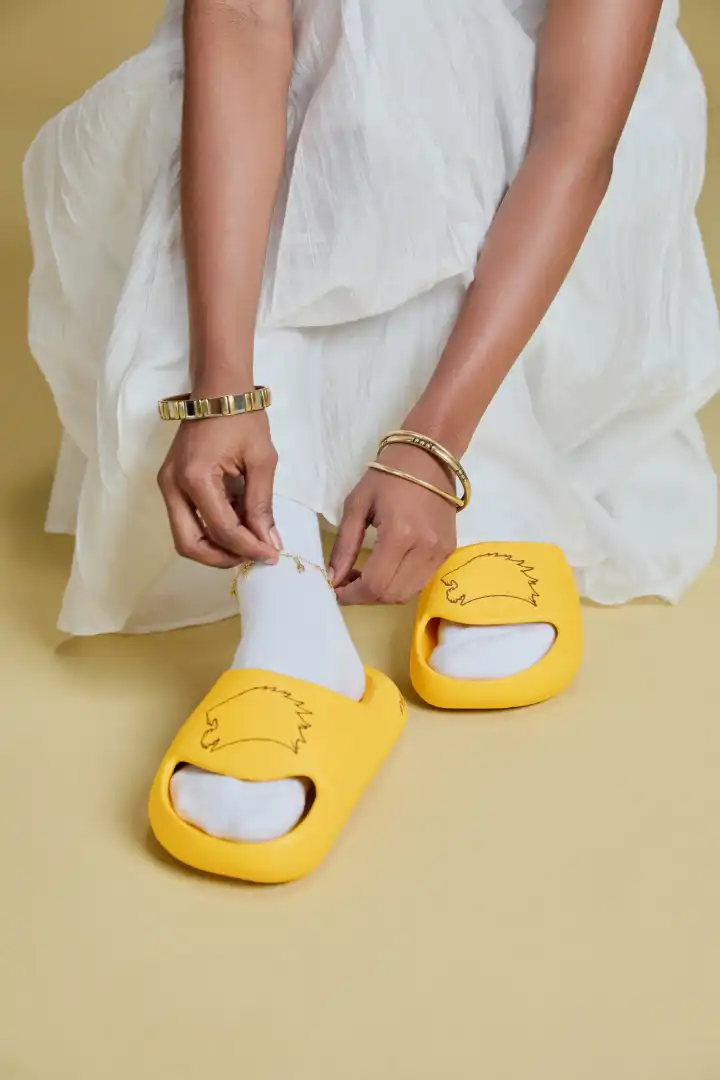 Person wearing white socks with yellow slides featuring a hedgehog design, fastening a gold anklet while dressed in a white outfit.