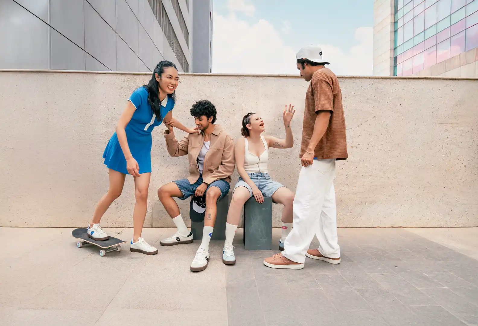 Four young adults socializing outdoors with one woman on a skateboard and two sitting on concrete blocks against a wall.
