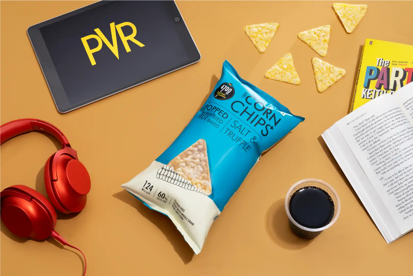 Bag of salted truffle corn chips surrounded by red headphones, tablet with PVR logo, book, coffee cup, and scattered chips on a tan surface.