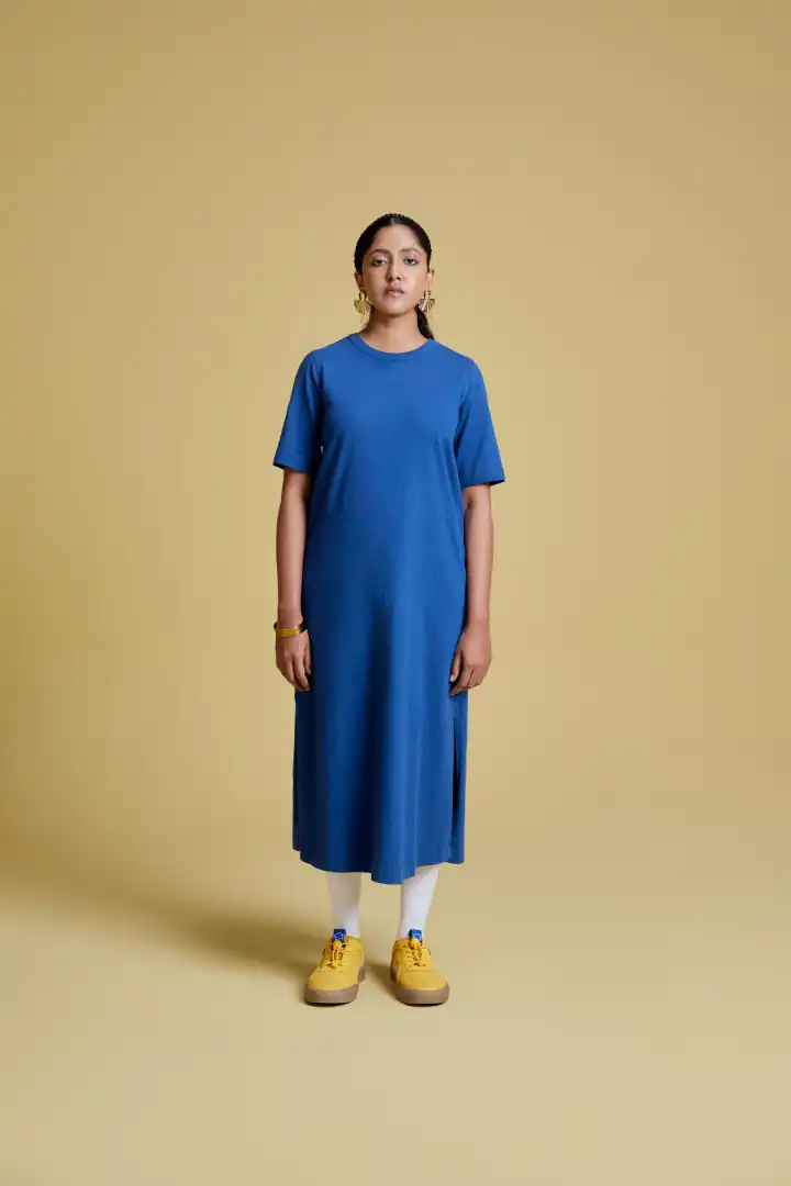 Woman standing against a beige background wearing a blue midi dress with short sleeves, white socks, yellow sneakers, and gold earrings.