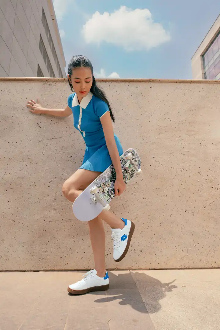 Young woman in a blue dress holding a skateboard, balanced on one leg against a beige wall outdoors under a blue sky with white clouds.