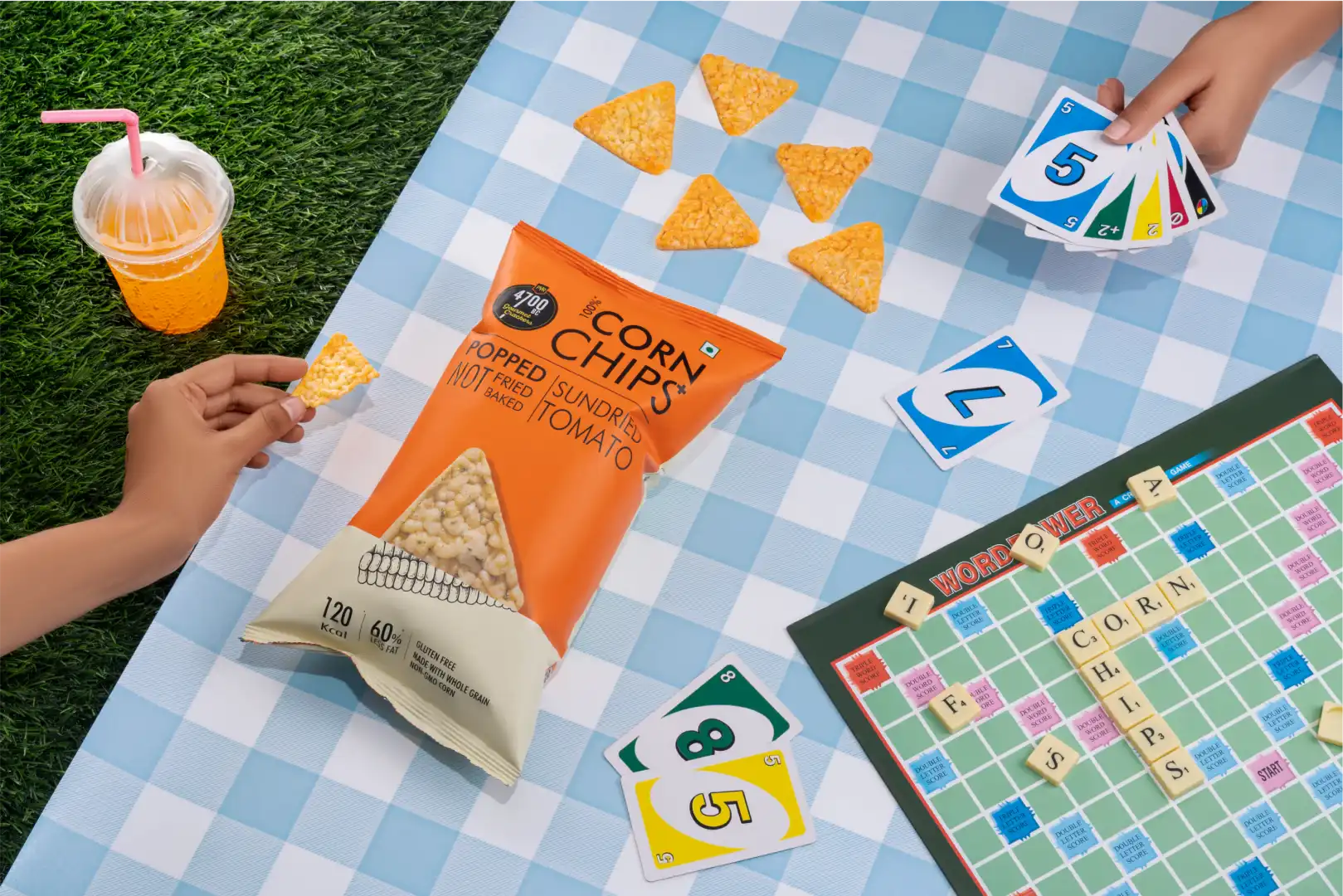 Picnic scene with a bag of tomato popped corn chips, scattered chips, a drink with a pink straw, UNO cards, and a Scrabble board spelling out 'CORN CHIPS'.