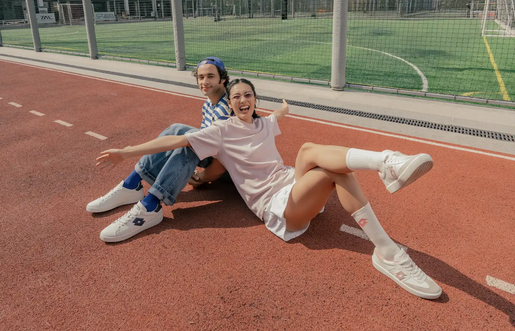Smiling young woman in light pink outfit and a young man in blue striped shirt and cap sitting on a red sports track with a green field behind them.