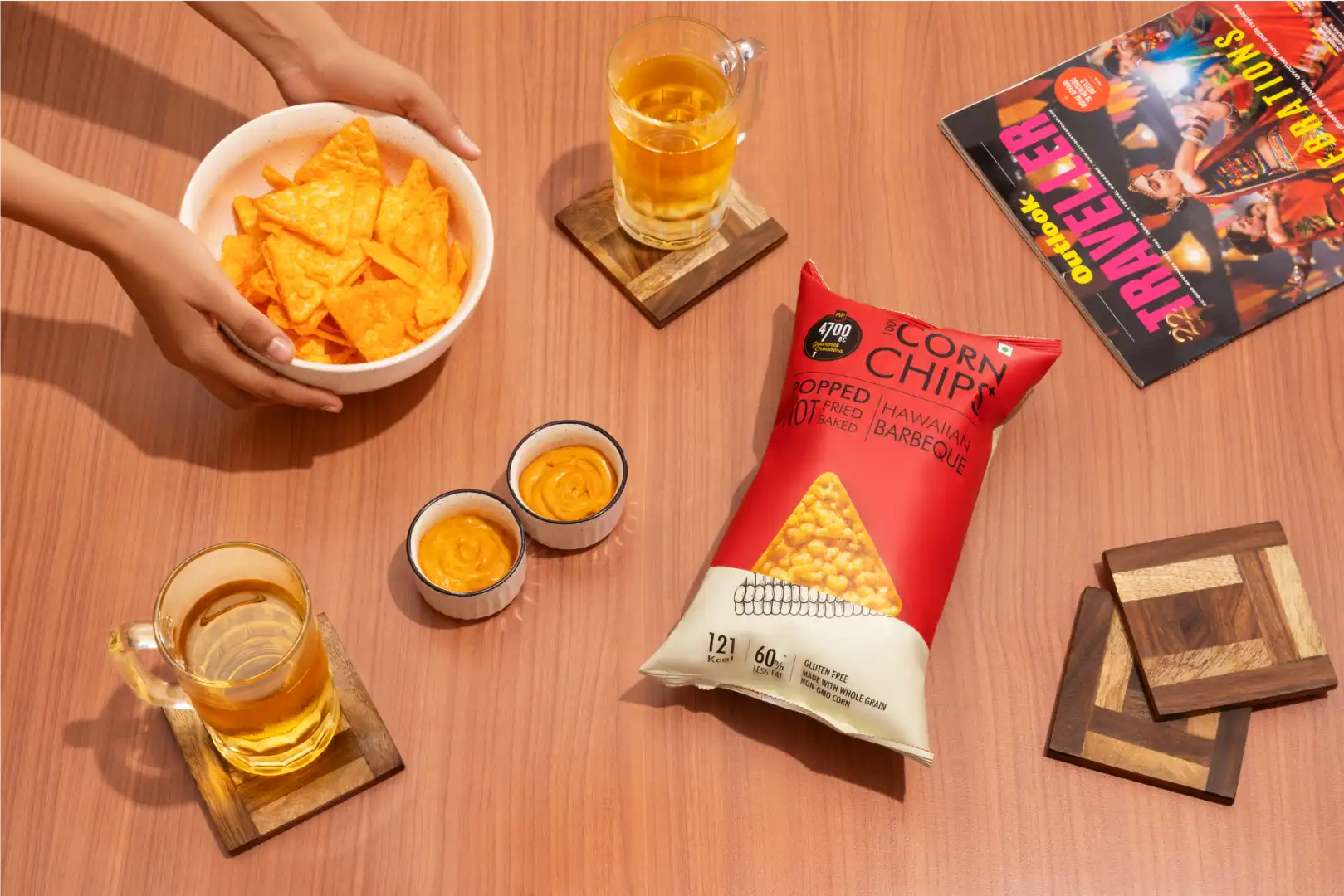Hands holding a bowl of orange corn chips on a wooden table with two dips, two glasses of light-colored drink on coasters, a red bag of Hawaiian Barbeque corn chips, and a magazine.