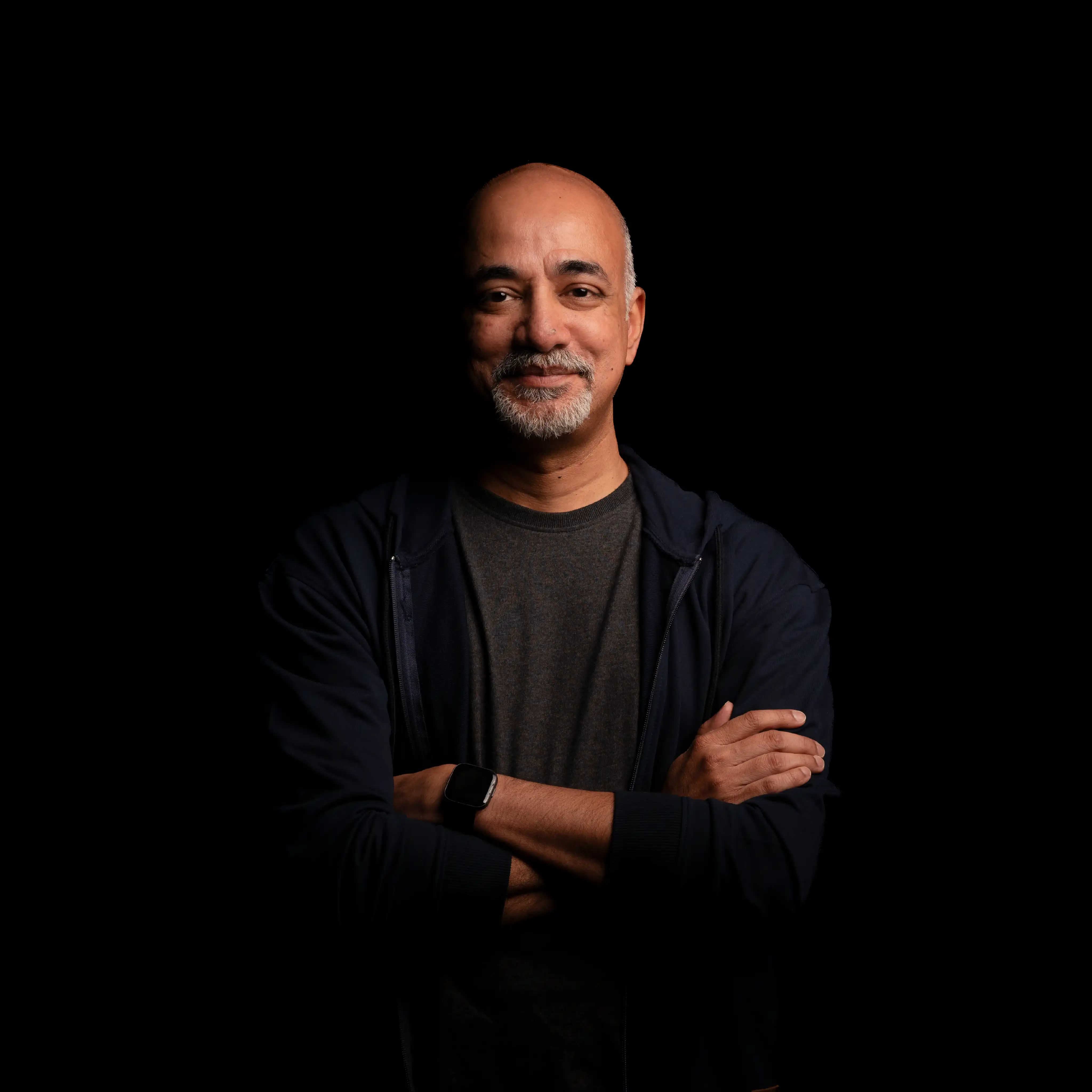 Man with a bald head and gray goatee wearing a black jacket and smartwatch, smiling with arms crossed against a black background.