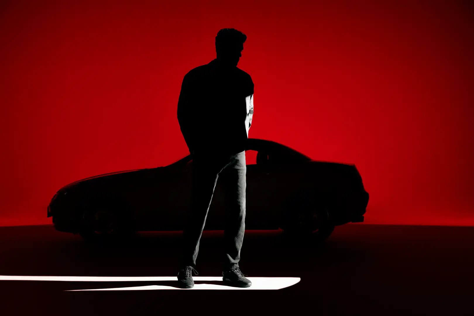 Silhouette of a man standing in front of a car against a red background with a spotlight on the floor.