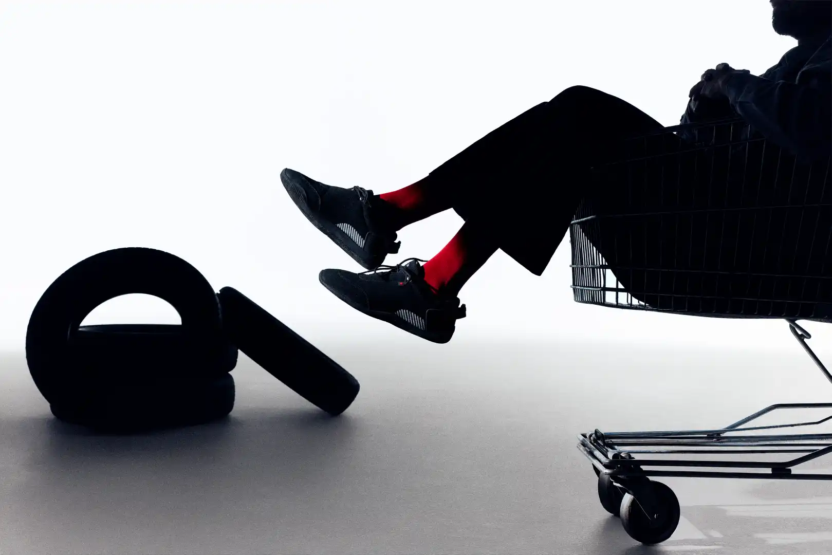 Silhouette of a person with red socks sitting in a shopping cart next to large black tires on a white background.