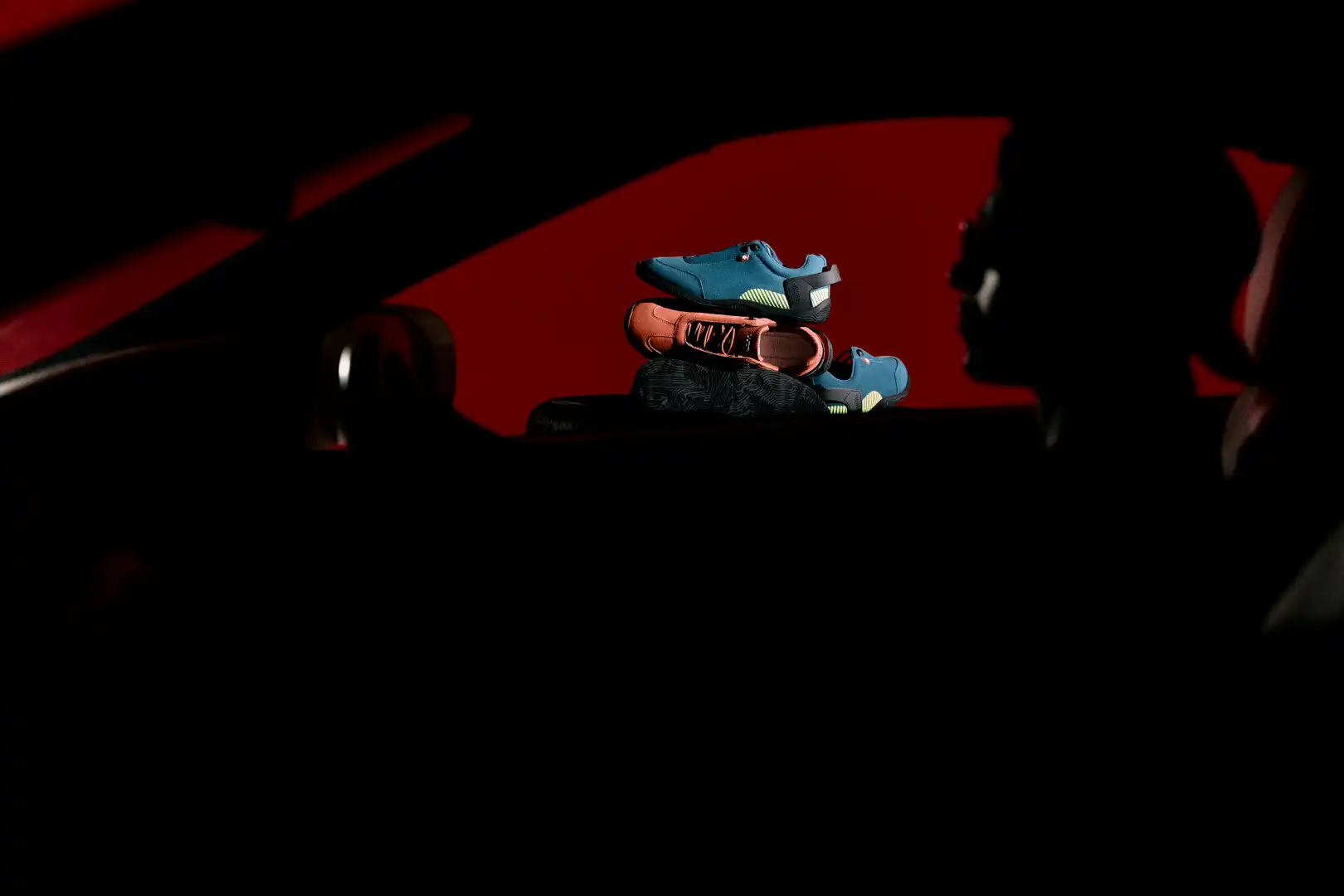 Stack of colorful athletic shoes on a dark surface with a person silhouette in a car against a red background.