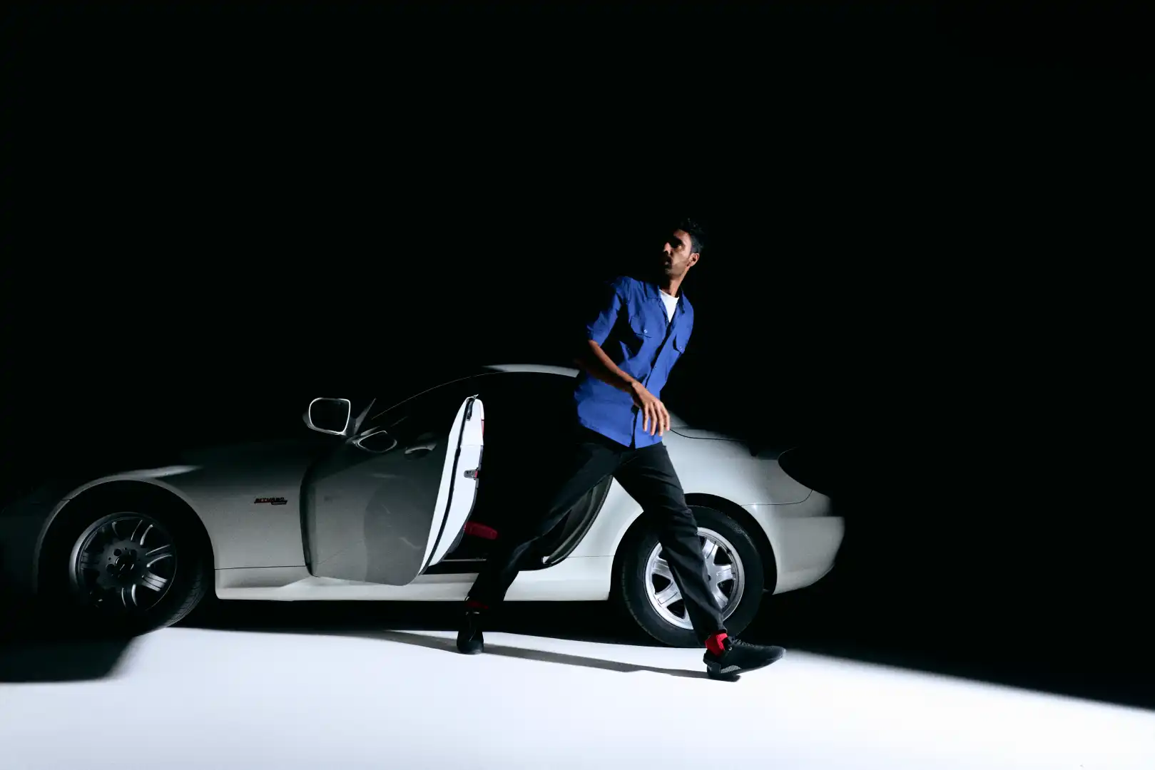 Man in a blue shirt stepping out of a white car with the door open, surrounded by darkness.