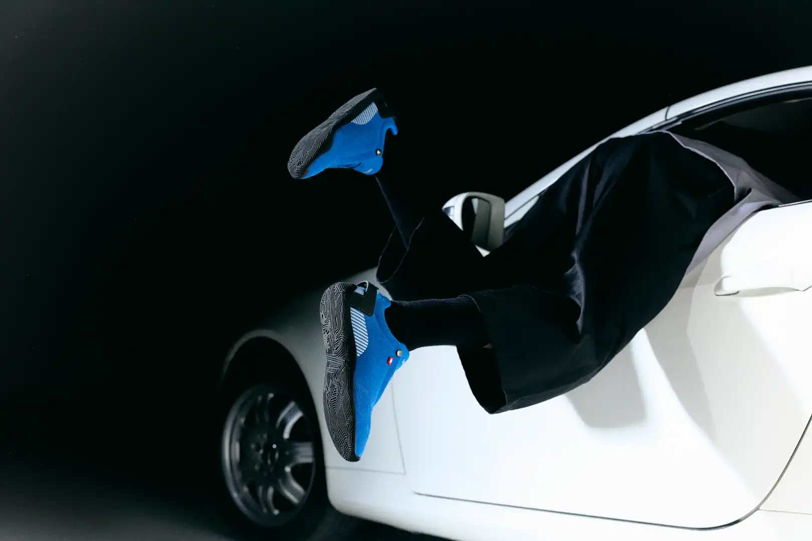 Person wearing black pants and blue sneakers with black soles stepping out of a white car at night.