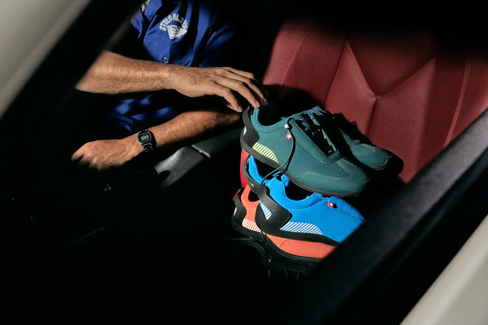 Stack of colorful sneakers including teal, blue, and coral on a red car seat, with a person wearing a watch and blue shirt nearby.