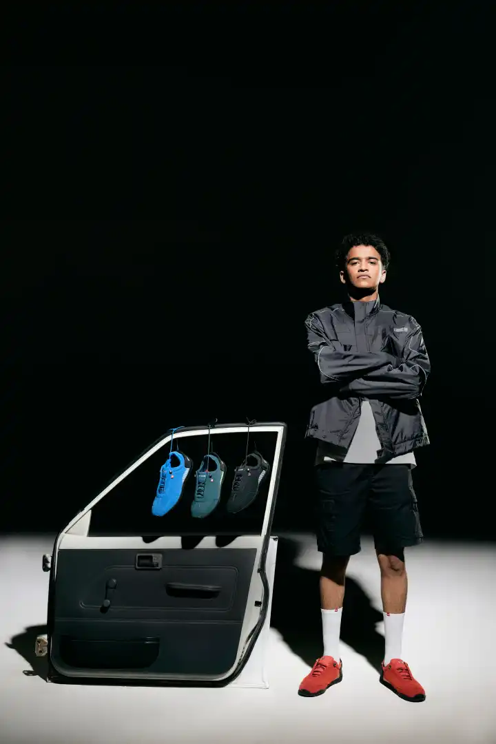 Man standing with crossed arms next to a car door hanging three pairs of shoes in blue, dark green, and black, wearing red sneakers.