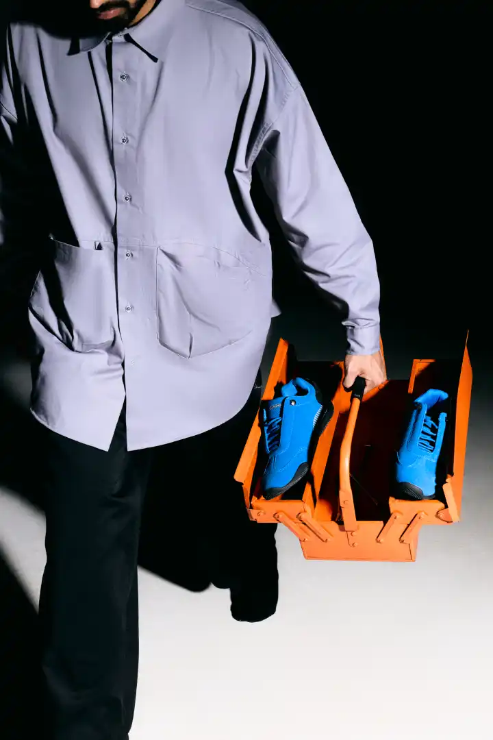 Person wearing a light gray shirt and black pants holding an orange toolbox containing bright blue shoes.