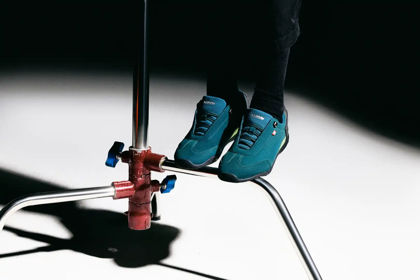 Pair of feet wearing teal Ludic sneakers standing on a metal pipe structure with a white and dark background.