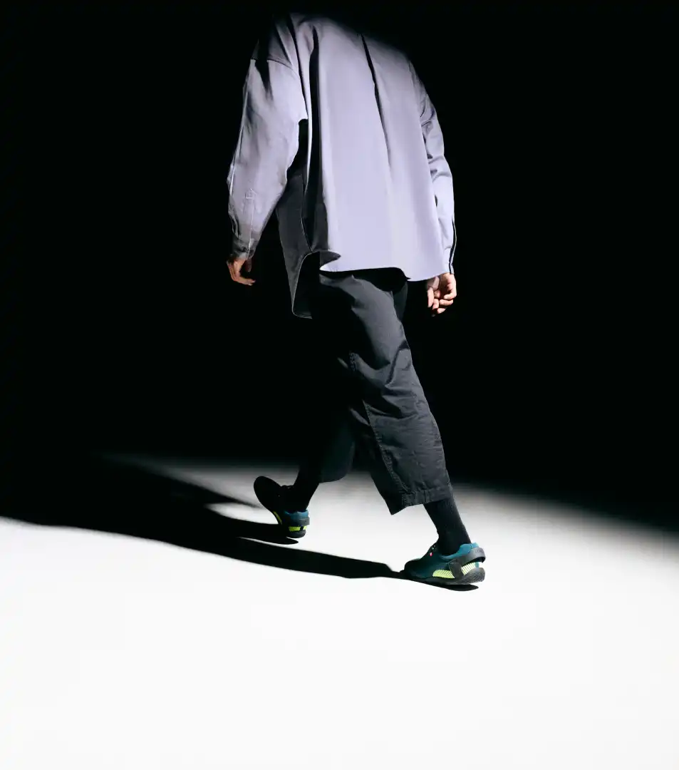 Person walking away wearing black cropped pants, black socks, gray shirt, and blue-green sneakers on a spotlighted floor with a dark background.