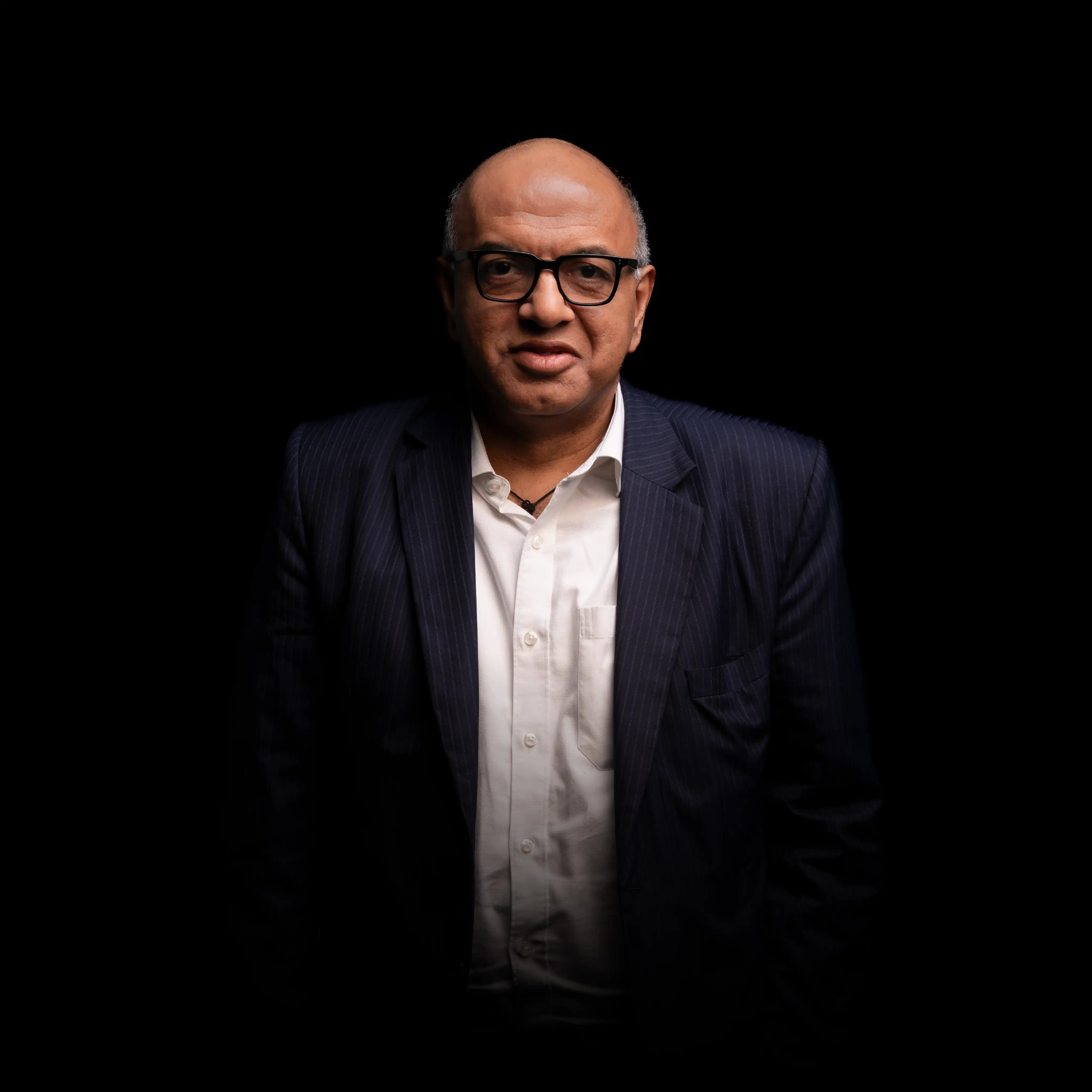 Portrait of a middle-aged man wearing glasses, a white shirt, and a dark pinstripe blazer against a black background.