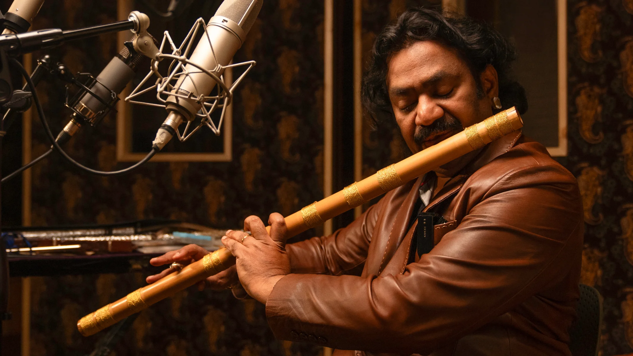 Man in brown leather jacket playing a decorated bamboo flute in a recording studio with microphones.