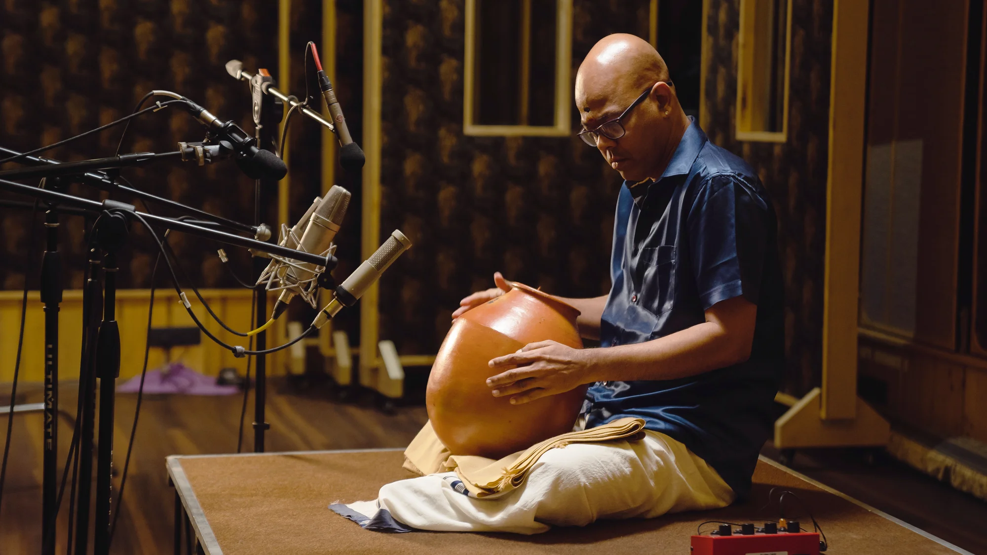 Man seated on floor playing a traditional clay pot drum in a recording studio with multiple microphones.